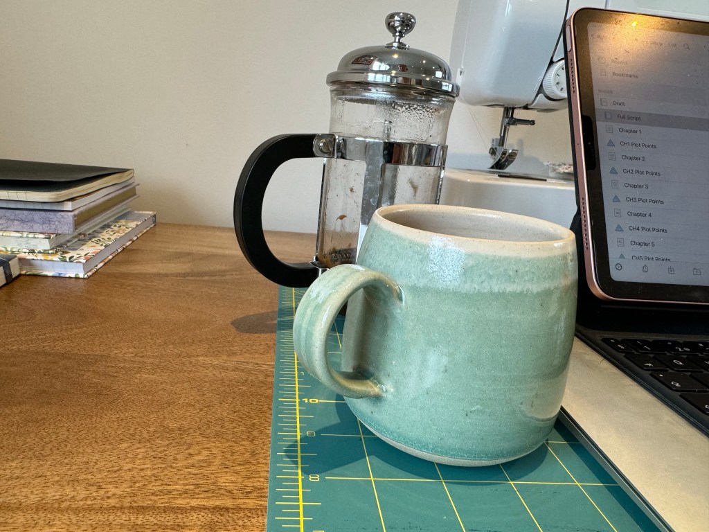 A desk with sewing, writing and coffee paraphernalia on it. 